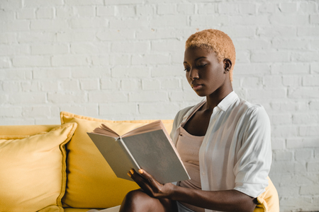 attractive african american woman with short hair reading book on yellow sofaの写真素材