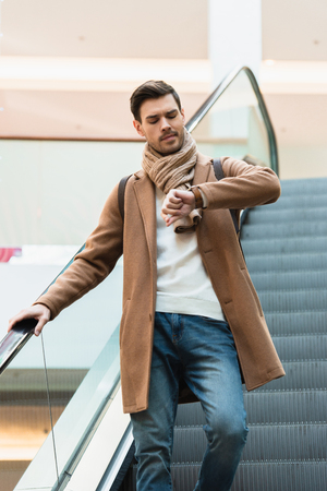 handsome man in going down on escalator and looking at watch in shopping mallの写真素材