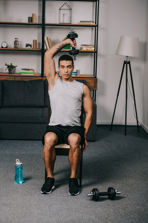 strong tattooed bi-racial man workout with dumbbell on chair in living roomの写真素材
