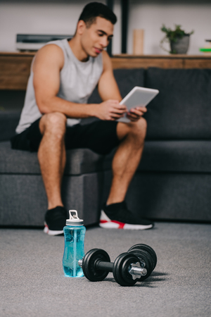selective focus of sport bottle near dumbbells with bi-racial man holding digital tablet on backgroundの写真素材