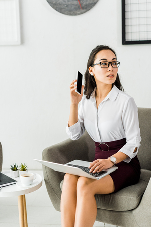 beautiful asian businesswoman sitting with laptop and using smartphone in officeの写真素材