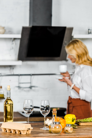 selective focus of middle aged woman holding cup of tea in kitchen, vegetables and wine on tabletopの写真素材