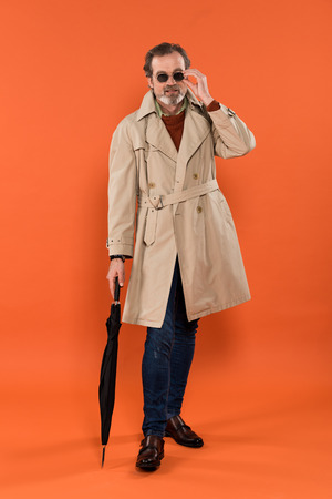 trendy senior man touching sunglasses and holding umbrella on orange backgroundの写真素材
