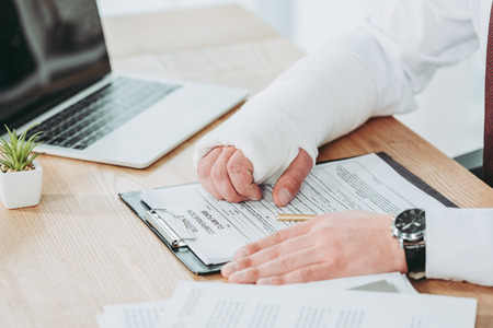 cropped view of worker with broken arm in gypsum sitting at table and holding hands on compensation form in office, compensation conceptの写真素材