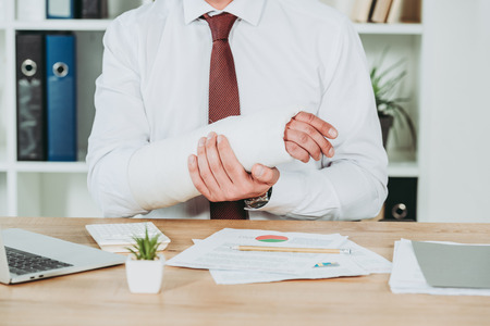 cropped view of worker holding broken arm wile sitting at table with documents in office, compensation conceptの写真素材