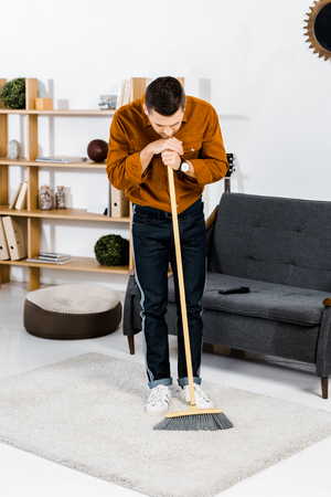 sad man standing with broom in modern living roomの写真素材