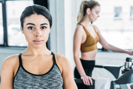beautiful african american sportswoman looking at camera and sporty girl exercising on treadmill behind in gymの写真素材