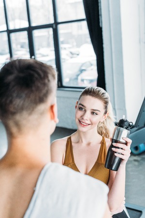 smiling young woman holding sports bottle and looking at athletic man with towel on foreground in gymの写真素材