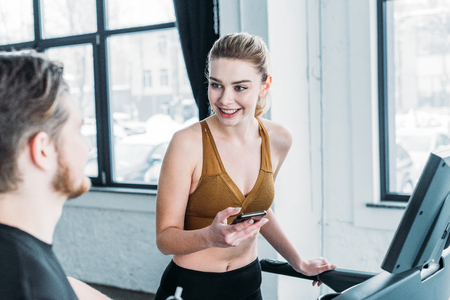smiling girl exercising on treadmill with smartphone in hand and looking at young man on foreground in gymの写真素材