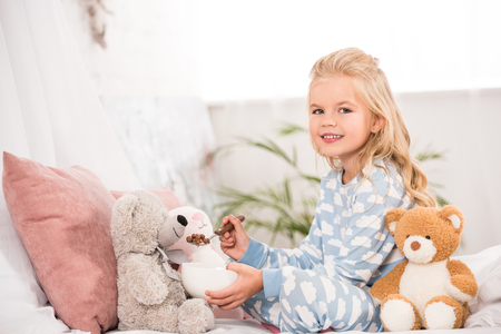 smiling child in pajamas feeding soft toys in bedの写真素材