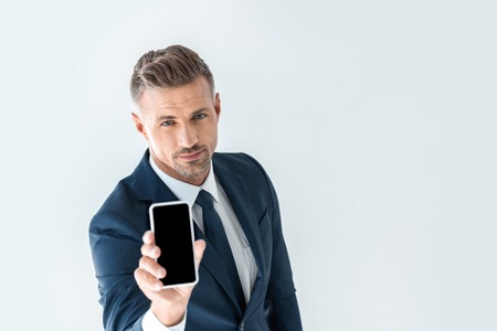 high angle view of handsome businessman showing smartphone with blank screen and looking at camera isolated on whiteの写真素材