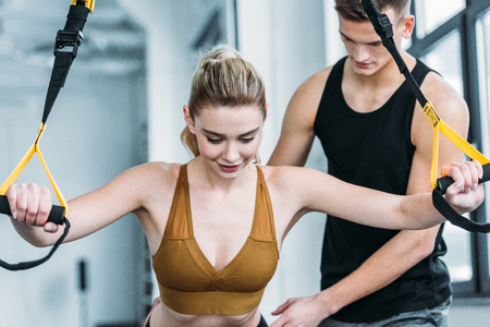 male trainer looking at smiling sporty girl exercising with suspension straps in gymの写真素材