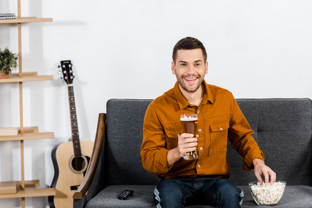 handsome man in modern living room holding glass of beer and eating popcornの写真素材