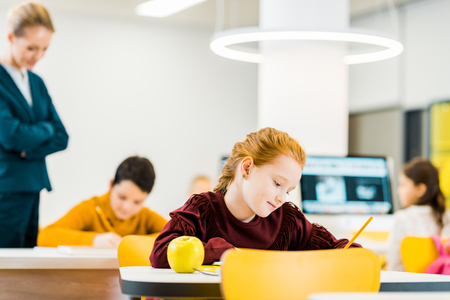 teacher looking at adorable schoolchildren writing and studying at desksの写真素材