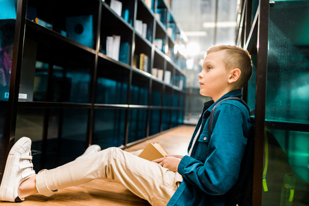 side view of cute schoolboy holding book and looking away while sitting on floor in libraryの写真素材