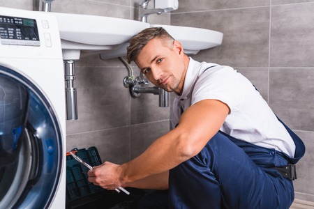 adult repairman sitting on floor and holding repair tool at bathroom and looking at camera