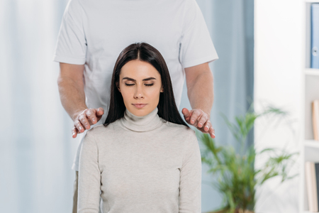 cropped shot of calm young woman with closed eyes receiving reiki healing treatment on shouldersの写真素材