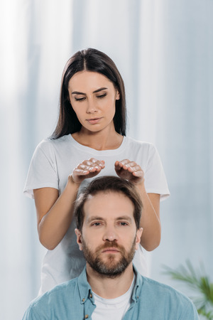 calm man looking at camera while receiving reiki treatment from young female healerの写真素材