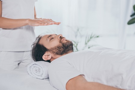 cropped shot of bearded man with closed eyes receiving reiki treatment above head