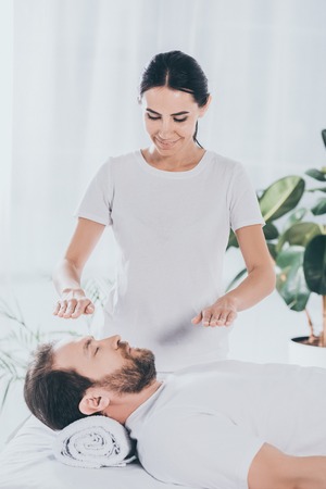 smiling young healer doing reiki session to calm bearded man with closed eyesの写真素材