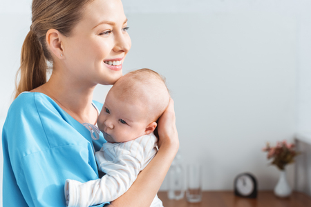 side view of happy young mother holding adorable baby and looking away in hospital roomの写真素材