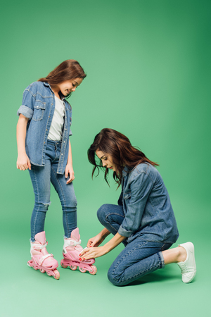 mother in denim putting roller blades on daughter on green backgroundの写真素材