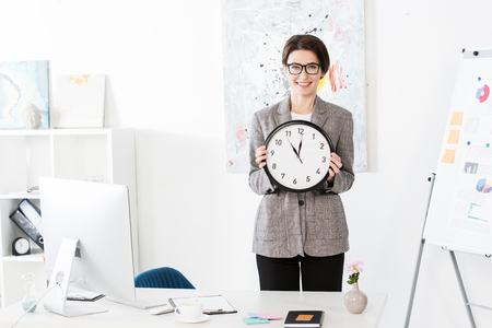smiling attractive businesswoman holding wall clock and looking at camera in officeの写真素材