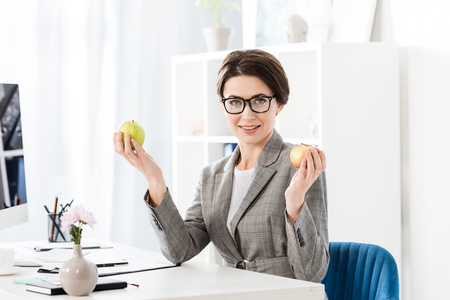 attractive businesswoman holding apples and looking at camera in officeの写真素材