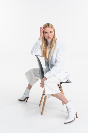 attractive caucasian blonde woman in fashionable white clothes sitting on chair and looking at camera isolated on whiteの写真素材