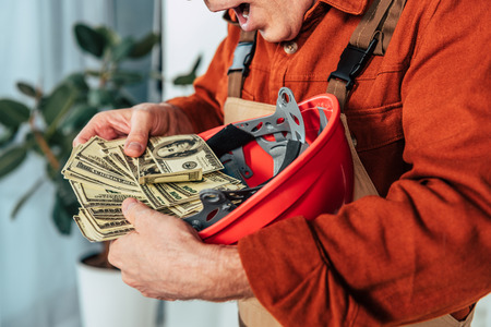 cropped view of surprised repairman holding hardhat with money in officeの写真素材