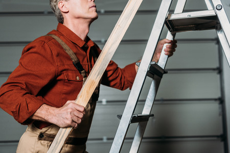 cropped view of repairman in orange uniform climbing with wooden board in hand on ladder in garageの写真素材
