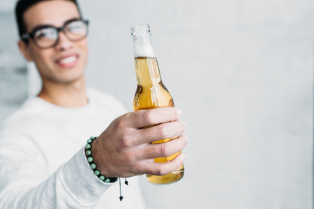 smiling mixed race man in glasses holding bottle of beer in stretched handの写真素材