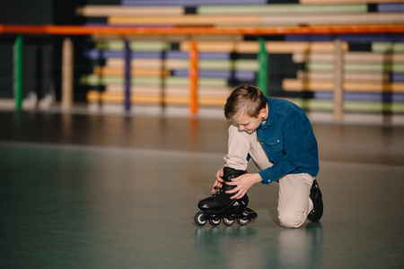 Selective view of cute boy sitting on floor and fixing roller skate bootの写真素材