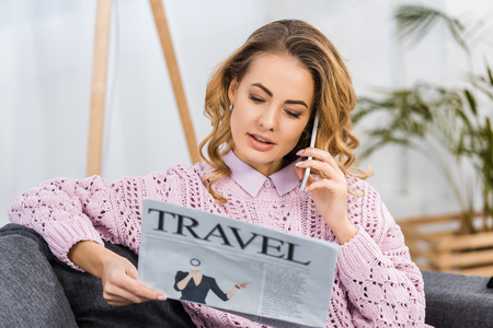 attractive woman sitting on sofa, talking on smartphone and reading travel newspaper in living roomの写真素材