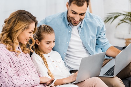 smiling parents sitting on sofa and looking at laptop holding by cute girlの写真素材