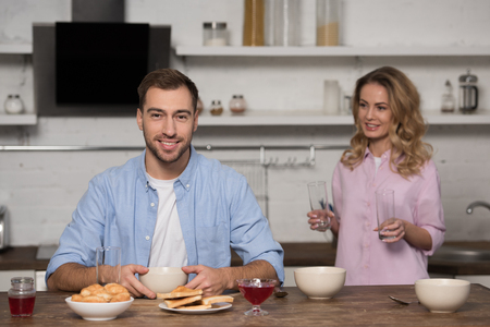 smiling man sitting at table and looking at camera with pretty wife holding glasses at backgroundの写真素材
