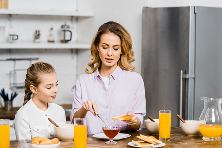 pretty woman spreading jam on toast for daughter in kitchenの写真素材