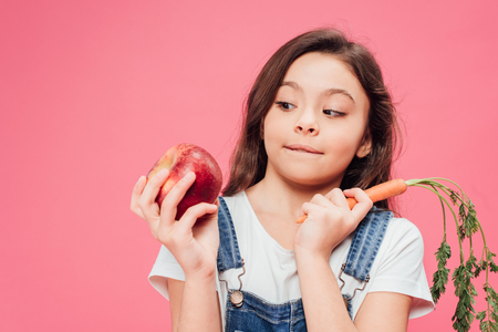 child looking at red apple and holding carrot isolated on pinkの写真素材