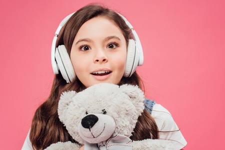 excited child in headphones holding teddy bear isolated on pinkの写真素材