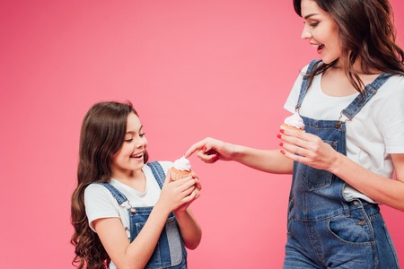 mother touching sweet cream on cupcake of daughter isolated on pinkの写真素材