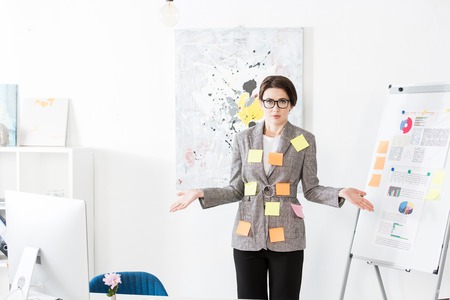 attractive businesswoman with paper stickers on jacket standing near flipchart and showing shrug gesture in officeの写真素材