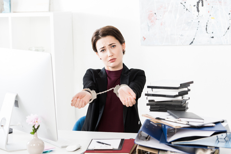 sad businesswoman showing hands with handcuffs at table in officeの写真素材