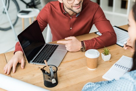 cropped view of colleagues sitting at table and using laptop with blank screen in officeの写真素材