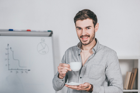 handsome young businessman holding cup of coffee and smiling at camera in officeの写真素材