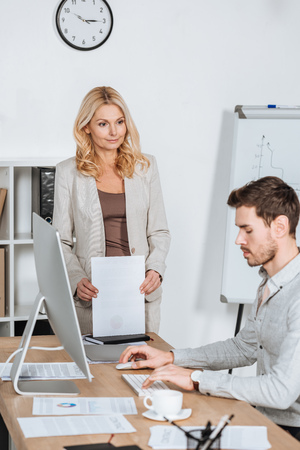 mature business mentor holding papers and looking at young businessman using desktop computer in officeの写真素材