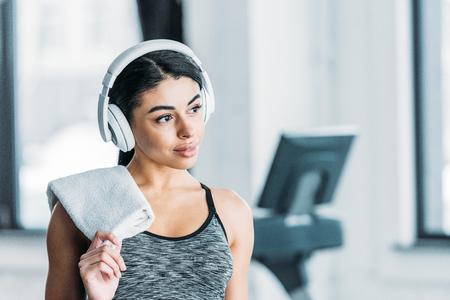 beautiful african american sportswoman in headphones holding towel and looking away in gymの写真素材