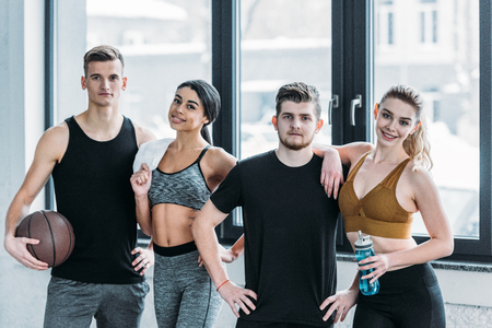 multiethnic group of sportive young friends standing together and smiling at camera in gymの写真素材