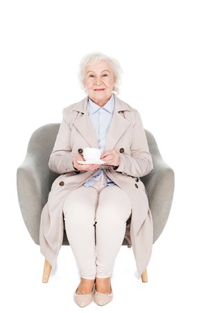 cheerful senior woman sitting in armchair with cup of tea isolated on whiteの写真素材