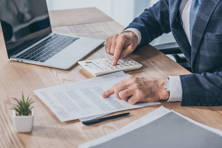 cropped view of businessman sitting at table, reading document and counting with calculator in office, compensation conceptの写真素材