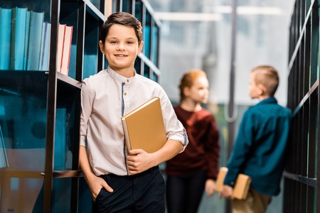 adorable boy holding book and smiling at camera while visiting library with classmatesの写真素材